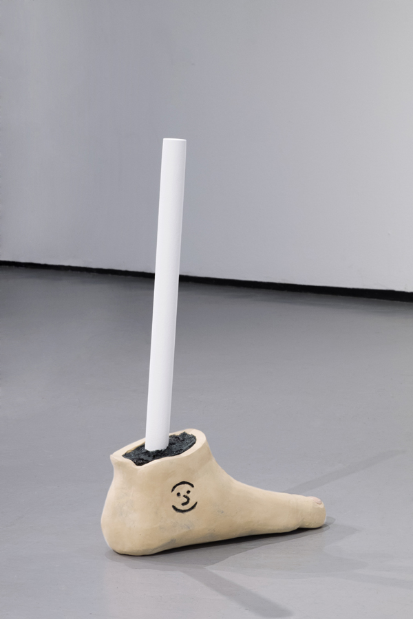 Closeup of a ceramic human foot sitting on the floor. The foot is cut at the ankle revealing a black foam interior core. A white cylindrical pipe emerges from the core like a bone into the air. A smiley face is etched into ankle of the foot like a tattoo.