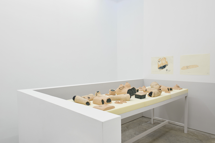 Installation view at the SculptureCenter. There is a long table covered in beige foam. Ontop lies various body parts made of painted ceramic. Arms, legs, torsos, chests, many of which also have tattoos carved into them in black. The walls of the room are bare white, except for two color drawings of some of the body parts on the table.