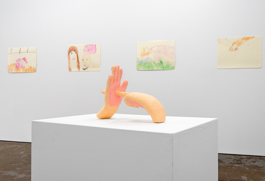 A sculpture of two peach-colored hands on a white pedestal. One hand is pointing its index finger through the palm of the other hand. There are four pen and crayon drawings on the wall in the background.