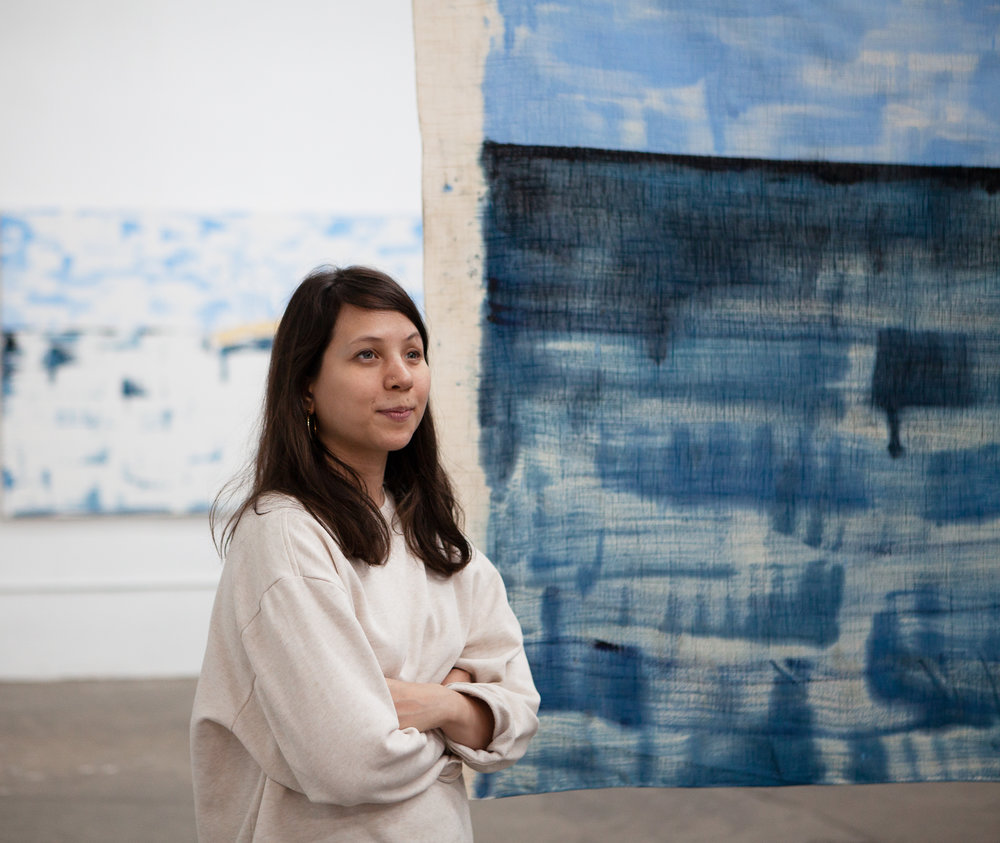 A portrait of Gossiaux standing in front of one of her paintings. She is wearing a beige sweater, crossing her arms, and striking a confident pose.