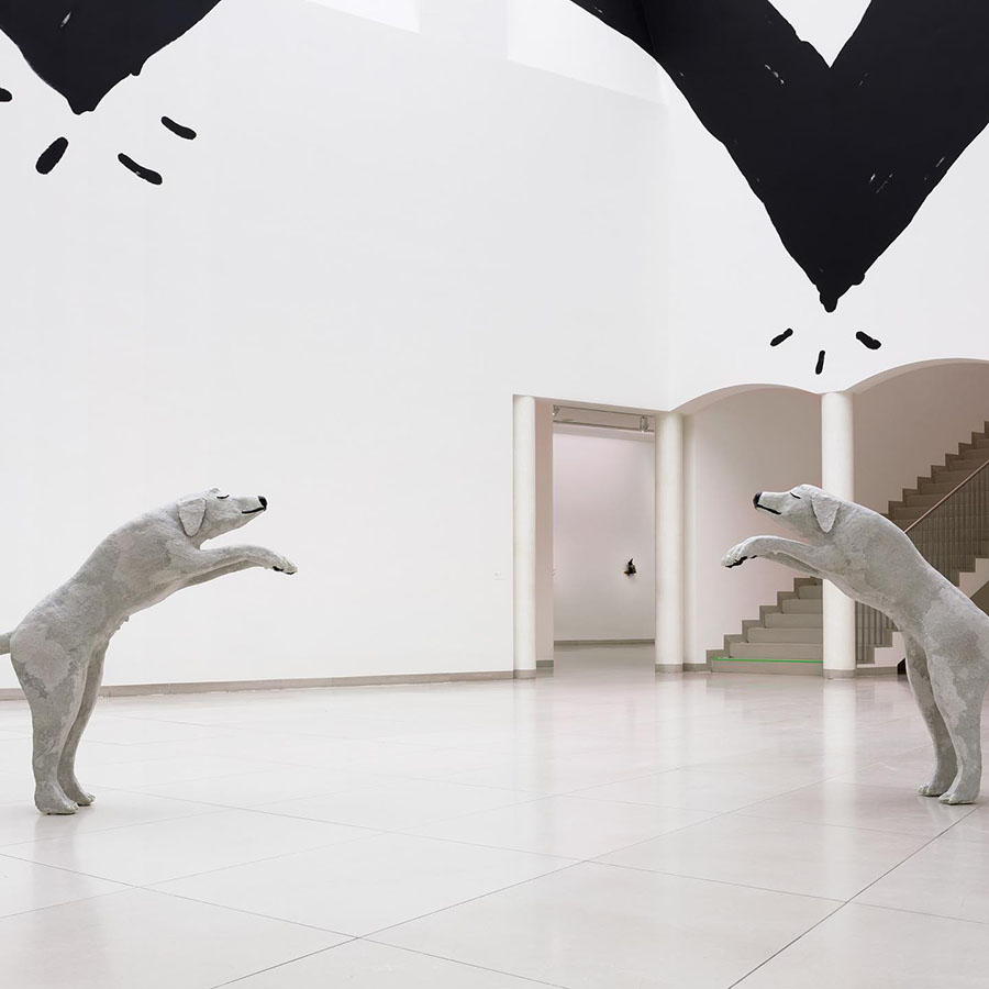 A photograph of MMK Frankfurt’s lobby, taken from above. On the floor below, two grey dog figures standing on their hind legs face each other. Behind them, a Christine Sun Kim painting stretches horizontally across the upper half of the 2+ story wall.