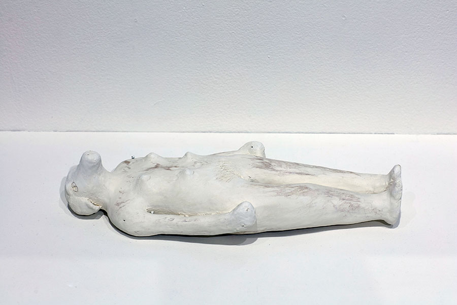 A doll sized white ceramic sculpture of an anthropomorphic dog with a woman’s body. The sculpture is resting on her back (on a shelf?), her hands are closed into fists, and are resting on either side of her hips. She has the head of a dog, with a long snout, and triangular shaped ears hanging down the side of her head, but with human features, such as her almond shaped eyes and long eyelashes, which are carved into the clay in thin lines. There are two small holes that were poked into the clay to create pupils in the center of each eye. On her torso she has four small breasts with nipples, a round belly with a belly button, and thin lines carved into the clay to signify pubic hair. She has long, straight legs with little round bumps to indicate knees, and her toes point up into the air, the back of her heels resting on the shelf.