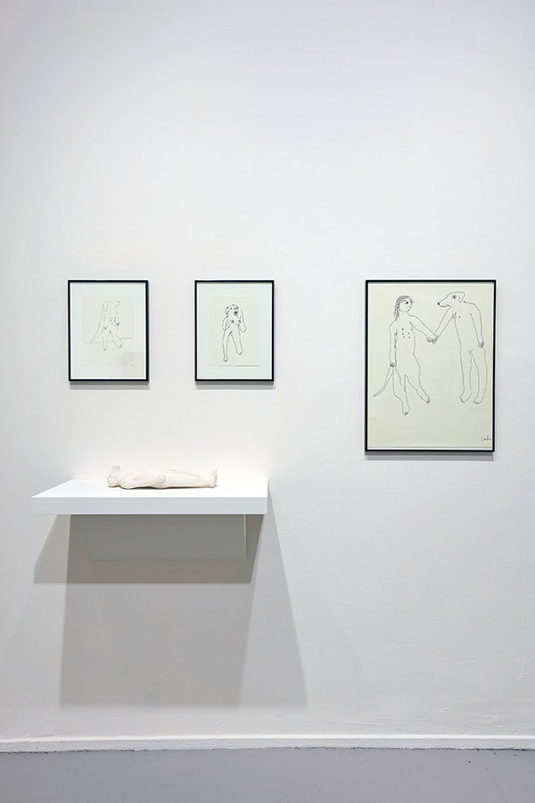 Hanging to the right of the 2 drawings is a simple line drawing, of two human animal hybrid figures. On the right side of the drawing, is a woman with a dog’s head, and on the left side is a dog’s body with a woman’s head. The two figures gaze affectionately at the other, as their bodies turn slightly inwards to face each other. The woman’s open hand reaches out to hold on to the dog figure’s paw.