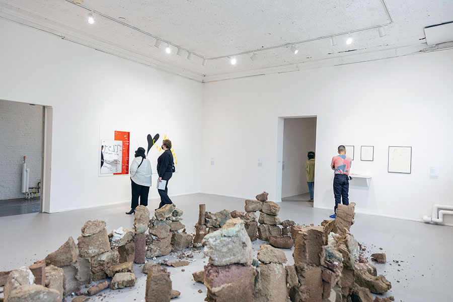 Installation view of large white gallery space with a crumbling stone sculpture in the center of the room and Gossiaux's pieces hanging on an adjacent wall.
