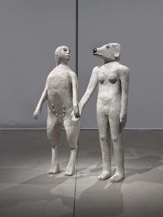 Two sculptures depicting hybrid human-animal figures stand side by side in a gallery space. They hold hands, with their heads slightly turned to gaze at one another. On the right, is a naked woman with a dog’s head; the figure on the left is an anthropomorphized dog with a long tail standing on its hind legs with the head of a woman.