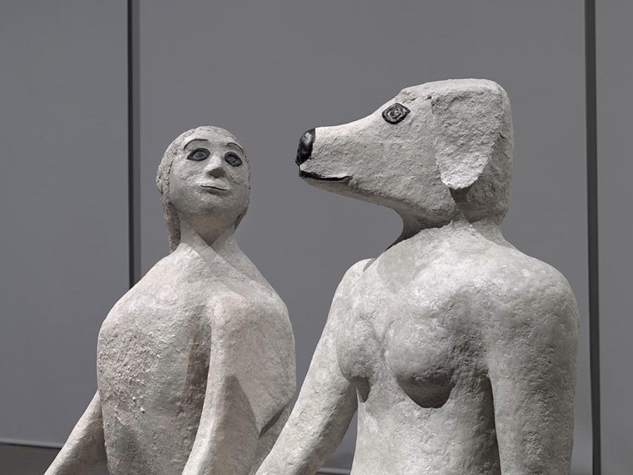 Two sculptures depicting hybrid human-animal figures stand side by side in a gallery space. They are seen close up, from the shoulders up, and from a slight profile view. On the right, is a naked woman with a dog’s head; the figure on the left is an anthropomorphized dog with the head of a woman.