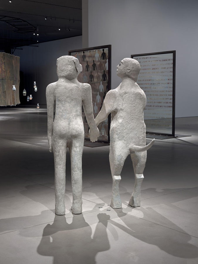 Two sculptures depicting hybrid human-animal figures stand side by side in a gallery space. They hold hands, with their heads slightly turned to gaze at one another, and are seen from behind. On the left, is a naked woman with a dog’s head; the figure on the right is an anthropomorphized dog with a long tail protruding, standing on its hind legs with the head of a woman. In the background are other sculptures and installations in the gallery.