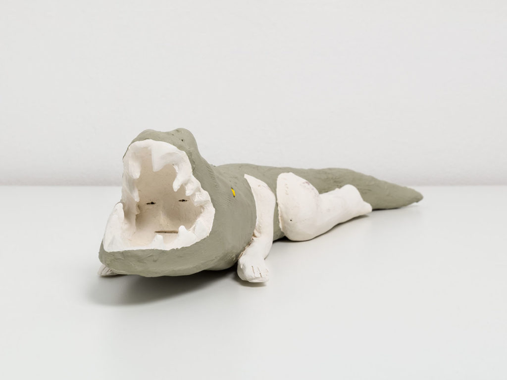 A ceramic sculpture of a humanoid alligator with woman’s legs and arms. The body of the alligator is painted jade green, and her almond shaped eyes are painted bright yellow. The arms and legs are left unpainted, and are the off white color of the clay. Her ams and legs are bent underneath her body, like she is crouching low, and her mouth is wide open with long jagged fangs. Inside of the alligator’s mouth is a person’s face with a nonplussed expression.