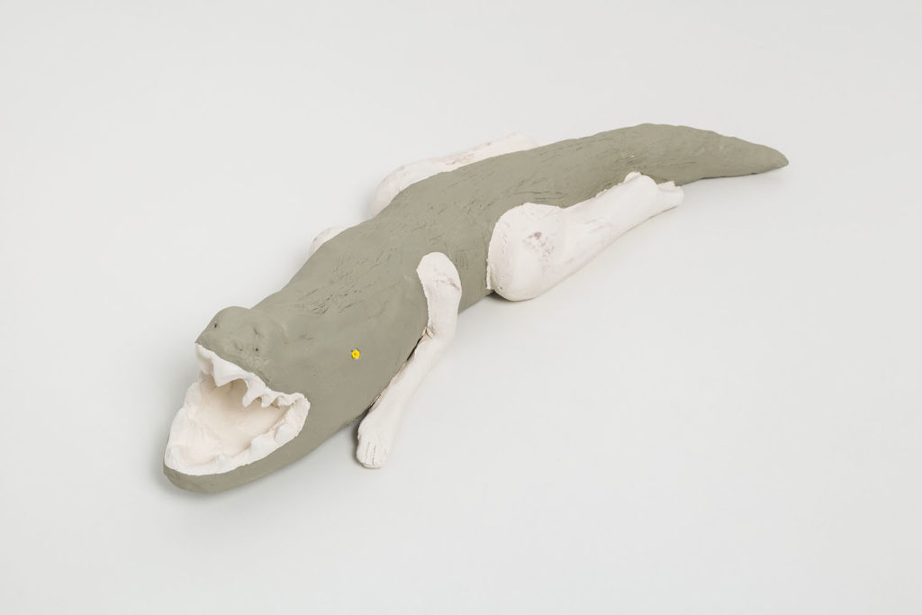 A ceramic sculpture of a humanoid alligator with woman’s legs and arms. The body of the alligator is painted jade green, and her almond shaped eyes are painted bright yellow. The arms and legs are left unpainted, and are the off white color of the clay. Her ams and legs are bent underneath her body, like she is crouching low, and her mouth is wide open with long jagged fangs. Inside of the alligator’s mouth is a person’s face with a nonplussed expression.