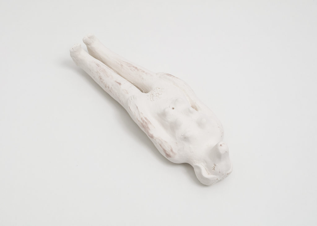 An off white doll sized ceramic sculpture of a woman with a dog’s head, and dog nipples lying down on her back. Her eyes are carved out from the clay to look like closed eyes with long eyelashes. Her left hand is resting on her left thigh, and her right hand rests flat against her right leg.