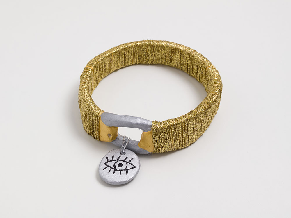 A ceramic sculpture of a dog collar hangs on the wall. The collar is wrapped with shiny gold string, and the dog tag is round, and painted silver with a black outline of a human eye with long eyelashes painted in the center.