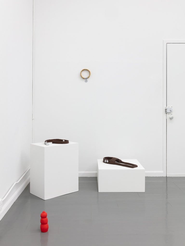 Installation detail view showing sculptures of a dog Leash and Harness displayed on low-height pedestals, a Red Kong sculpture on the floor, and a Golden Dog Collar hung on the wall.