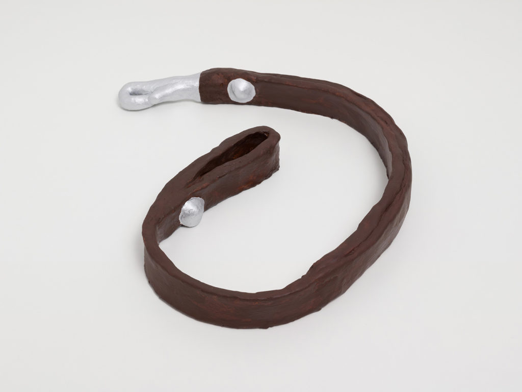 A ceramic sculpture of a dog leash wrapped up in a loose coil. The ceramic is painted chocolate brown to look like soft leather, and the metal clasp and buttons are painted silver.