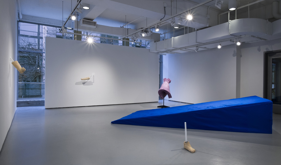 Installation view of a pristine whitewall gallery space. A large cobalt blue wedge made of fabric and foam in the middle of the gallery. Sculptures of body parts are installed sparsely on the walls and placed on the floor.