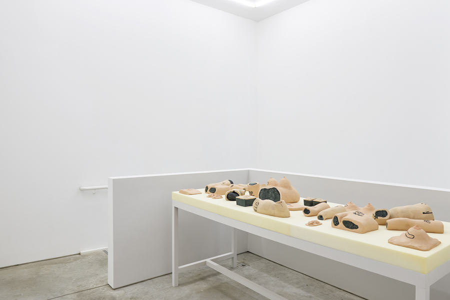 Installation view at the SculptureCenter. There is a long table covered in beige foam. Ontop lies various body parts made of painted ceramic. Arms, legs, torsos, chests, many of which also have tattoos carved into them in black. The walls of the room are bare white, except for two color drawings of some of the body parts on the table.