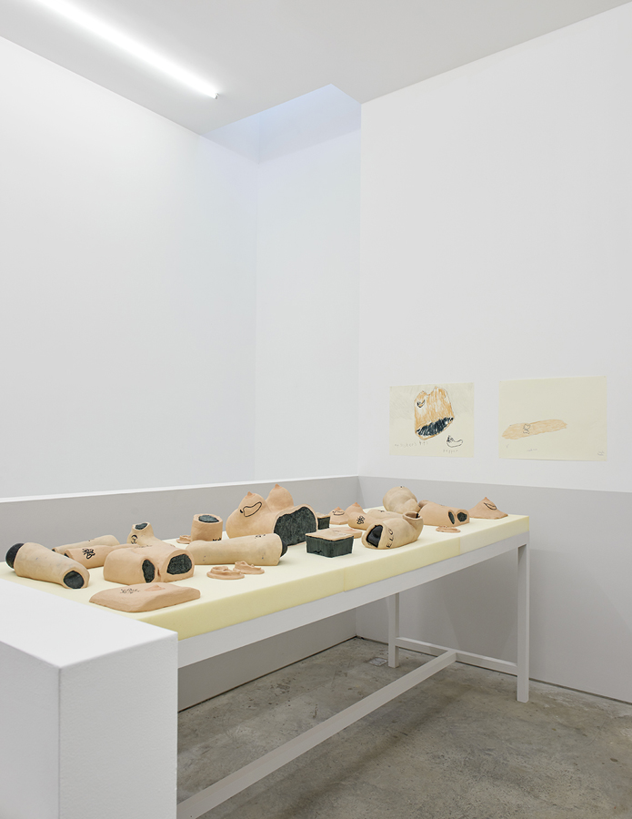 Installation view at the SculptureCenter. There is a long table covered in beige foam. Ontop lies various body parts made of painted ceramic. Arms, legs, torsos, chests, many of which also have tattoos carved into them in black. The walls of the room are bare white, except for two color drawings of some of the body parts on the table.