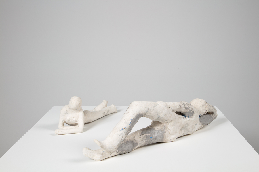 Two small white papier mache figures on a pedestal, facing each other. A female figure lies on her stomach while a male figure lies on his side.