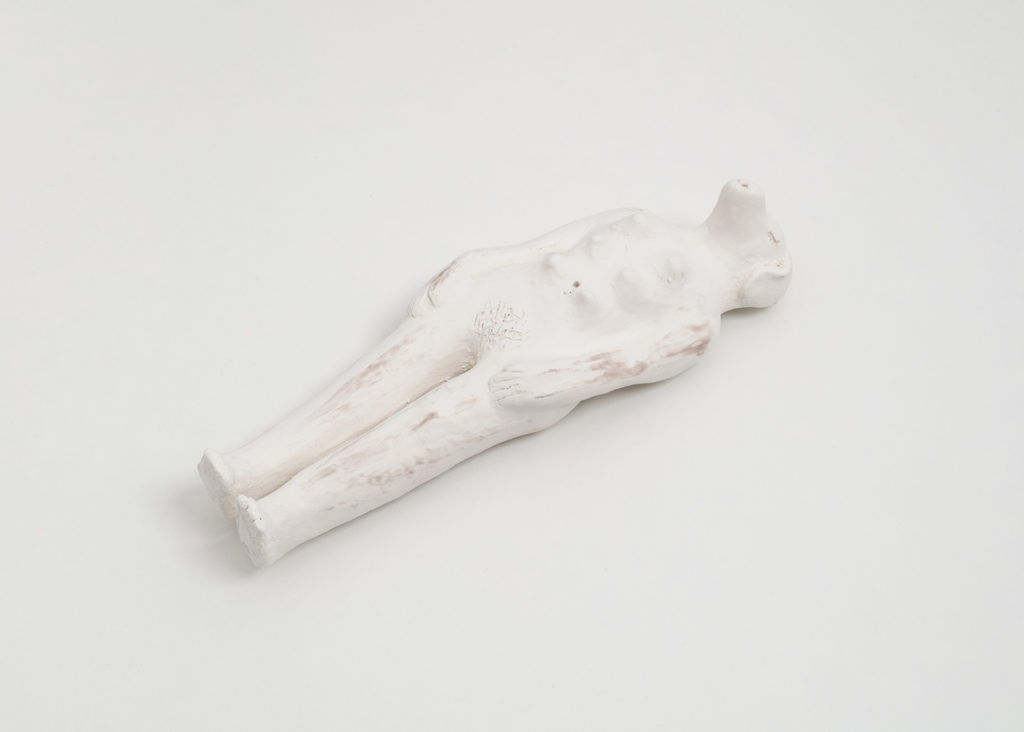 An off white doll sized ceramic sculpture of a woman with a dog’s head, and dog nipples lying down on her back. Her eyes are carved out from the clay to look like closed eyes with long eyelashes. Her left hand is resting on her left thigh, and her right hand rests flat against her right leg.