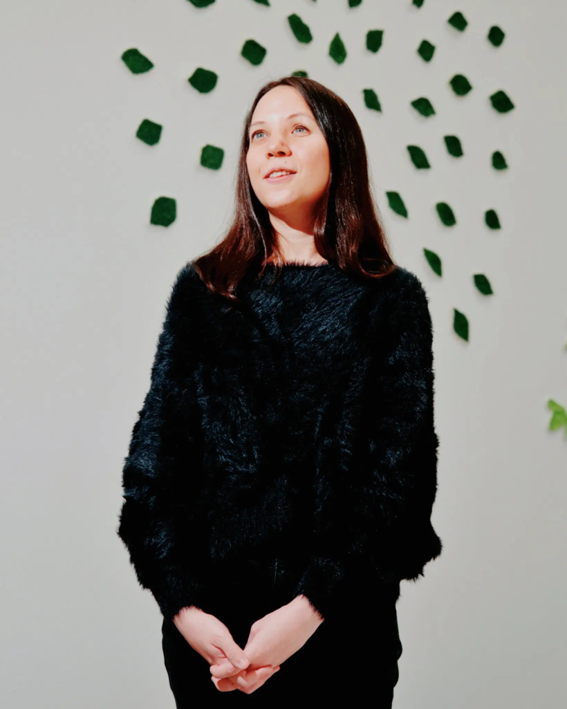 A beatific smile on the face of Emilie Gossiaux, seen at the Queens Museum, in a gallery where textured papier-mâché leaves she made are affixed to a wall to suggest a tree.