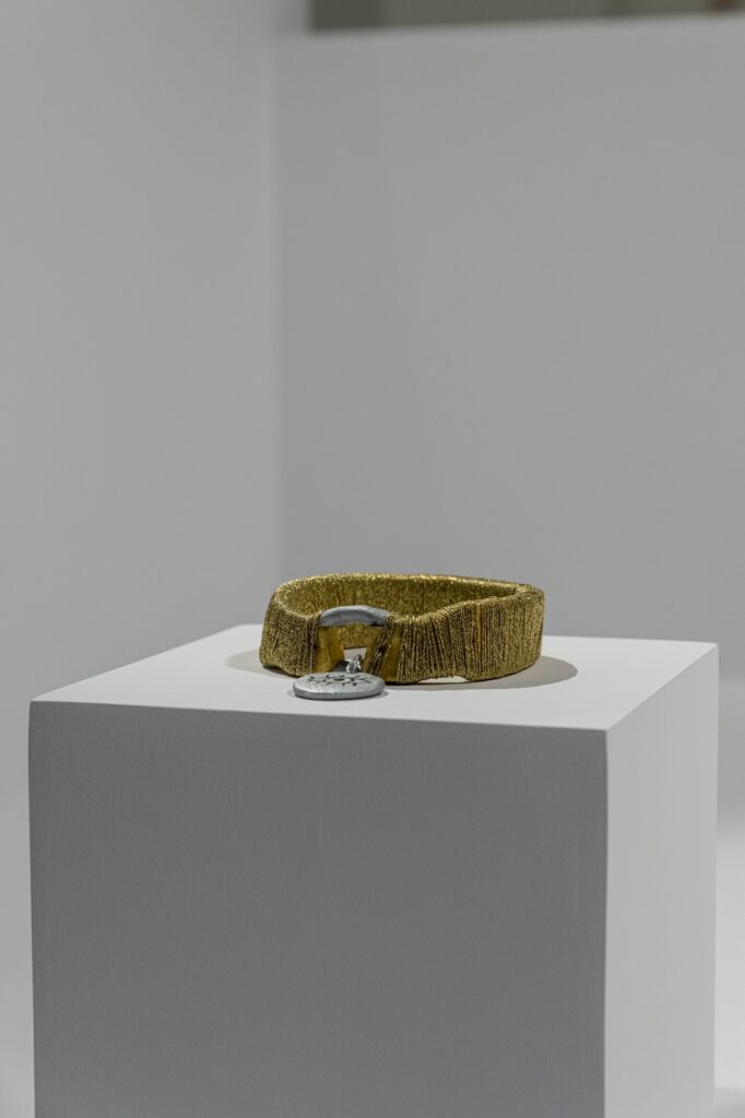 “Golden Dog Collar” is a circular ceramic sculpture made to resemble a dog collar. The main loop has been painted a metallic gold and there is a silver buckle at the front. Reflective gold colored string has been wrapped tightly around the gold painted part of the collar. Hanging from the buckle is a flat ceramic disk painted in metallic silver. The disc is a little larger than a quarter. A line drawing style eye has been painted on the face of this silver disc in black.