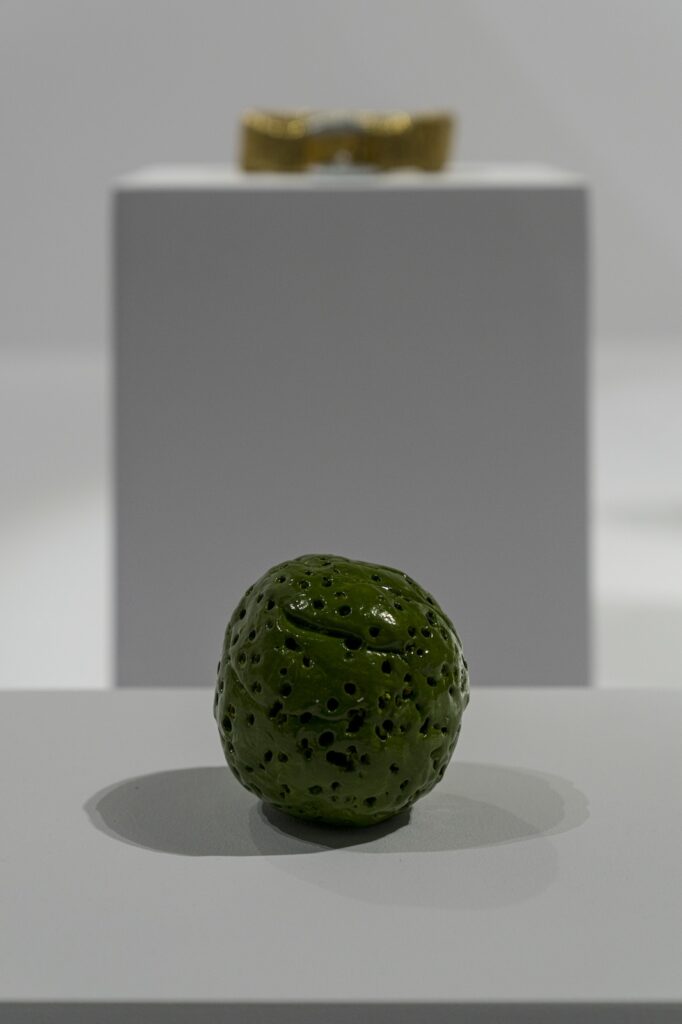 "Green Ball" is a small ceramic sculpture in the shape of a lopsided ball. It is a little larger than a baseball. Small, irregularly sized holes have been pricked in the surface and it is painted a shiny yellow-green. In this vertical installation shot, "Golden Dog Collar" sits on a separate plinth behind, slightly out of focus.