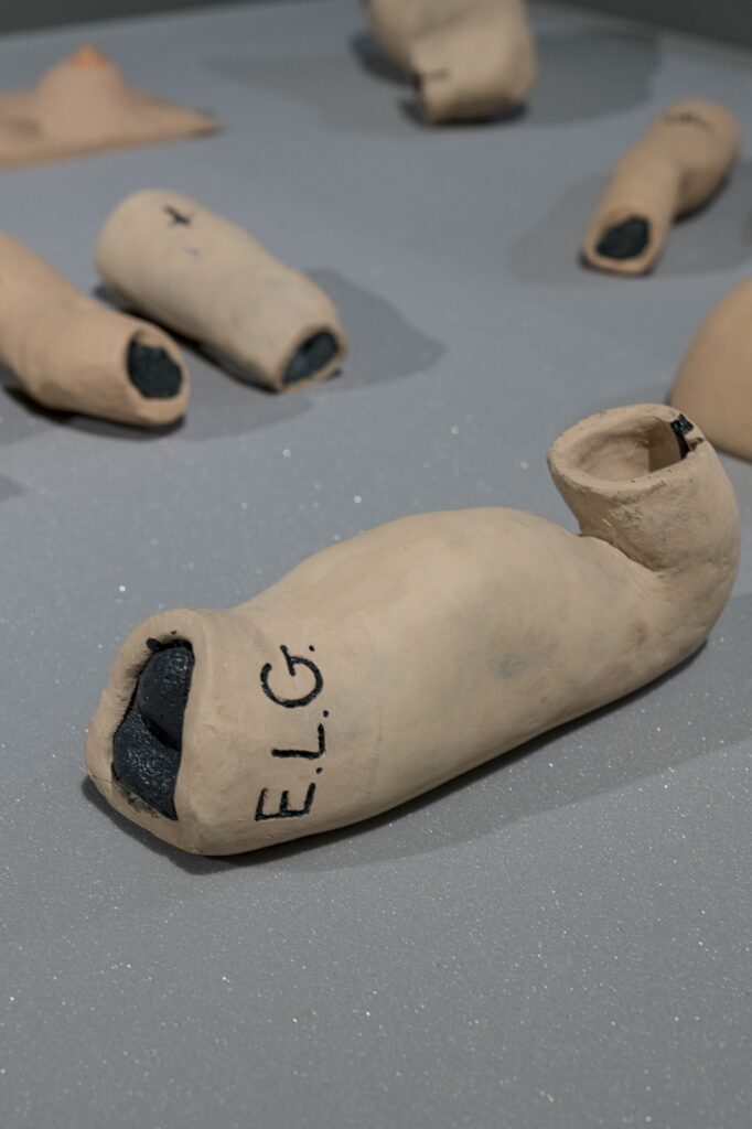In this vertical installation shot of "E.L.G. Familial Archives”, the image is focused on a flexing bicep with the initials 'E.L.G.' applied to its side. Behind the bicep, three forearms, another bicep, and a pair of breasts are visible but out of focus.