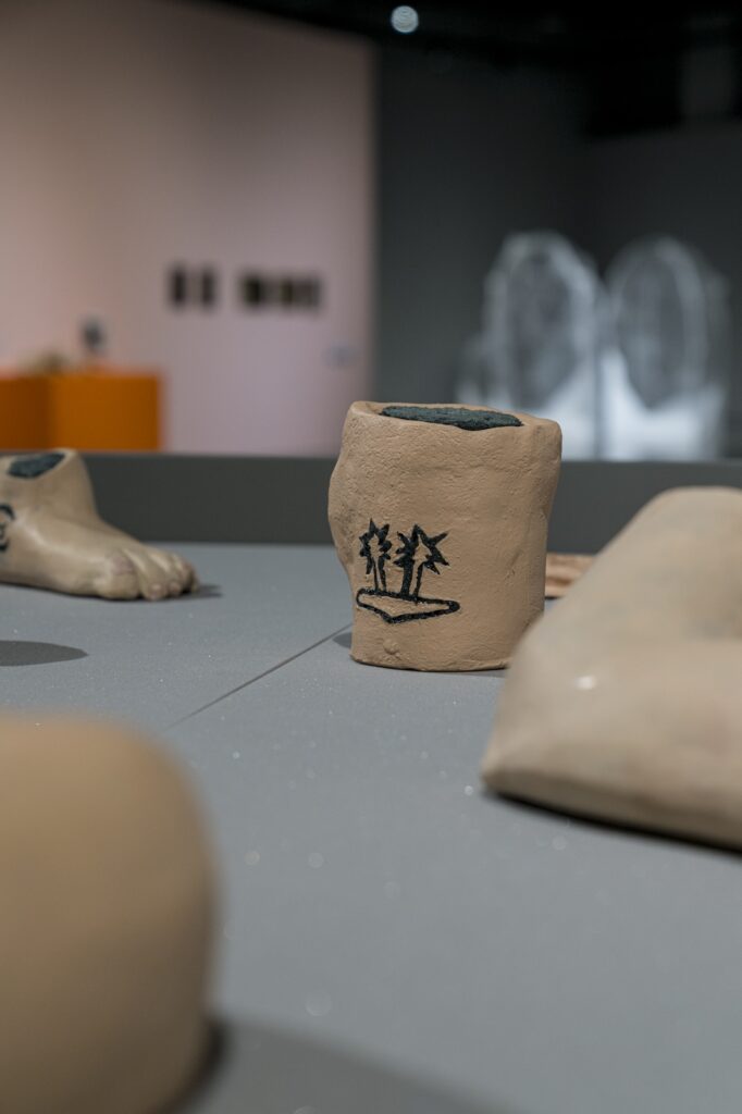 In this vertical installation shot of "E.L.G. Familial Archives”, a sculpture resembling a neck with the line drawing of a desert island with a pair of palm trees is the focus. To the left, slightly out of focus, the toes of a foot peek into the frame.