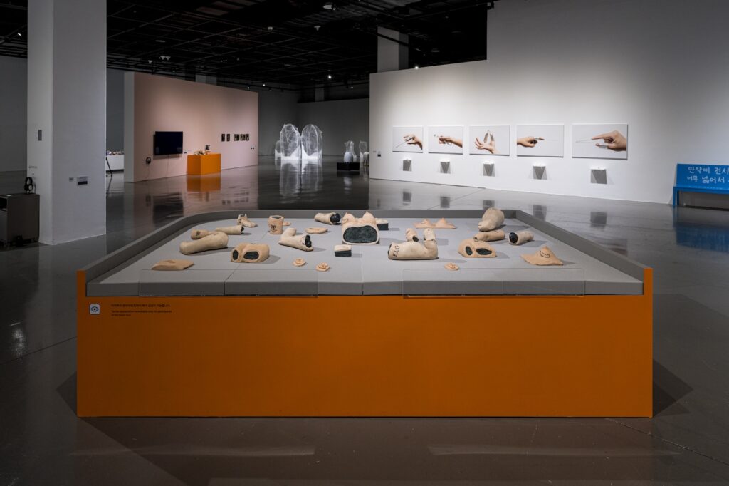 In this installation shot, the entirety of "E.L.G. Familial Archives” is spread across the foam topped table. From this angle, the orange painted sides of the table are visible, so are various pieces of art by other artists in the background.