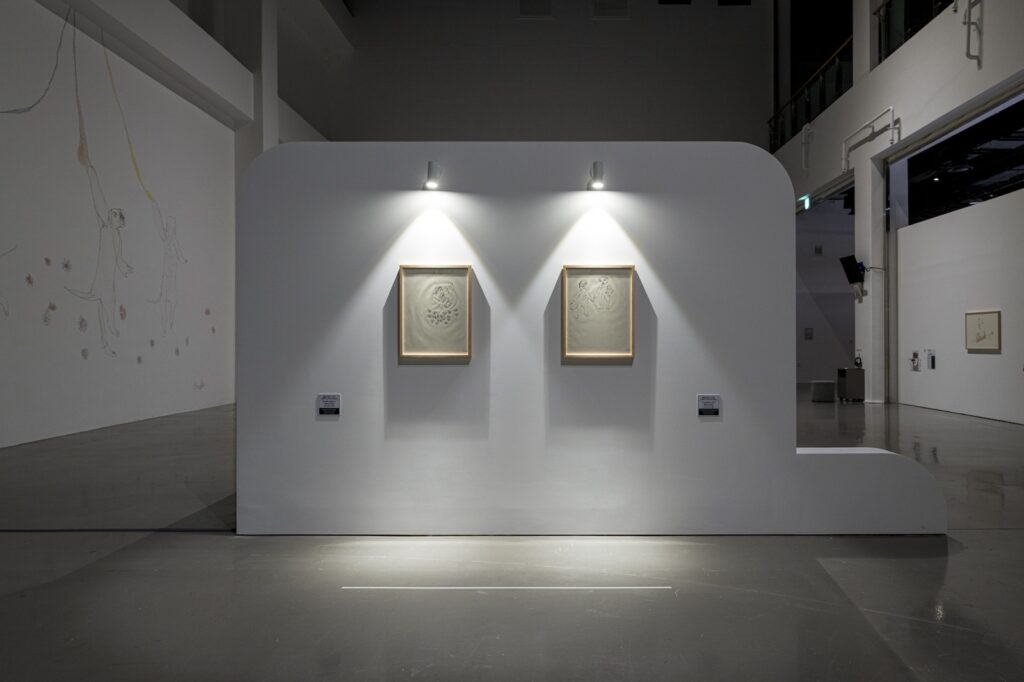 In this vertical installation shot from "Seeing With 10 Fingers", two drawings, "Return Under the Moon” and “Hybrid Animals Holding Hands” are hung side by side on a freestanding white wall that has been set up in the middle of the gallery. There is a downward facing light installed above each drawing, illuminating the paper while the frame creates long shadows. The mural is visible to the left.