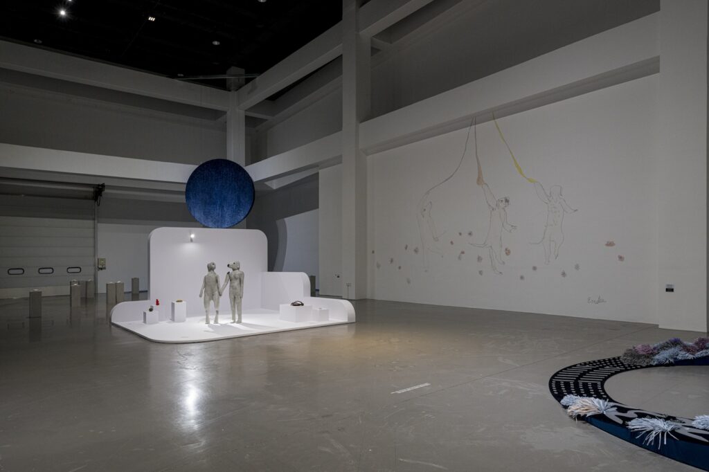 In this installation shot from "Seeing With 10 Fingers", a large mural, adapted from the drawing "Londons Dancing with Flowers", has been applied to the wall. In the mural, three English Labrador Retrievers stand on their hind legs, like they are in the middle of a dance. Each one holds onto a brightly colored leash that disappears into the ceiling above, from left to right, the leashes are colored purple, red, and yellow. Purple and red flowers are scattered around the dogs' feet. To the left of the mural, “True Love will Find You in the End” and the other sculptures have been installed on a freestanding platform.