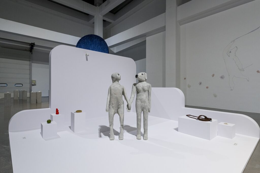 In this installation shot from "Seeing With 10 Fingers", the two hybrid human-animal figures from “True Love will Find You in the End” stand side by side in the middle of two groups of plinths. To their left, three plinths hold "Green Ball", "Kong", and "Golden Dog Collar". To their right, two plinths hold "Harness" and "Treats". Part of the mural is visible behind.