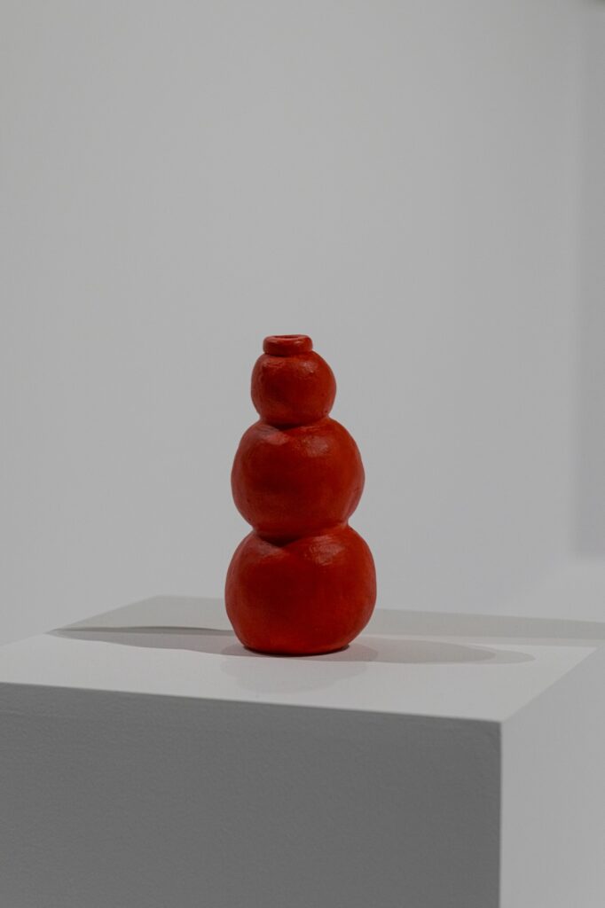 "Kong" is a cylindrical ceramic sculpture painted bright red. It is made up of three connected spheres, with the largest on the bottom and the smallest on top. The bottom of the biggest sphere is flat, enabling the sculpture to stand upright. There is a small, ridegelike ring on the top of the smallest sphere. This sculpture is reminiscent of the rubber kong toys that dogs play with.