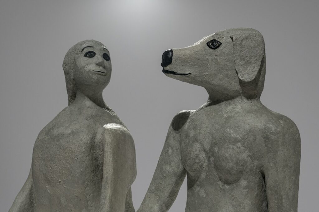 “True Love will Find You in the End” consists of two hybrid human-animal figures sculptures standing side by side in a gallery space. They hold hands, with their heads slightly turned to gaze at one another. The figure on the right is a naked woman with a dog’s head; the figure on the left is an anthropomorphized dog with a long tail standing on its hind legs with the head of a woman. The surface on both sculptures resembles concrete and the facial details are matte black.