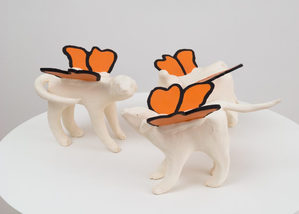 “Butterflydogs” consists of a trio of ceramic sculptures, each made to resemble a white English Labrador Retrievers. Their legs are thick and slightly uneven, giving a handmade, almost childlike appearance. Their tails are long and each one sticks out in a different direction, one straight back, another to the right in a 'u' shape, and the last drops straight down. On each dog's back are a pair of large, orange butterfly wings with black outlines, reminiscent of monarch butterflies. The wings are upright and attached at the center of the dogs’ backs, making them look as if they could up and fly off. The three figures are positioned close together, as if interacting or forming a small group.