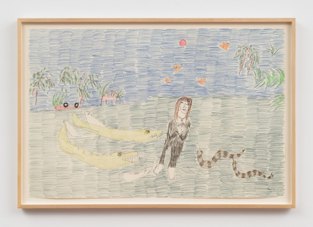“In My Dreams, The Mississippi Carries Me” is a colorful drawing on paper, framed in a light wood frame. The scene is set outdoors and is divided horizontally: the lower half depicts a dark green body of water, and the upper half is a sky filled with blue horizontal strokes. In the foreground, on the left side of the drawing, are two large, green alligators with woman arms and hands splashing out of the water. Their mouths are open, and their eyes are round and cartoonish, and their teeth are visible. The Alligatorgirls are swimming towards a woman standing barefoot in the center-right, who has pale skin, shoulder-length brown hair, and a wide, closed-mouth smile. She wears a black outfit that covers her arms and legs. Her hands are clasped together at her chest, and her posture is relaxed and peaceful. To the right of the woman, are two long, brown and black striped snakes with yellow eyes and long black eyelashes slithering in the water towards her. The sky is a vibrant blue, with a bright orange sun at the top center, and scattered in the sky are five orange butterflies. On the left side, at the water’s edge, there are three small, leafy trees with brown trunks and green leaves. Between the trees, there is a day bed with magenta sheets, and a red car that has been overturned with its black wheels in the air. On the far right side of the drawing, another leafy tree with long, thin green leaves is visible. The overall style is dreamlike and wild.