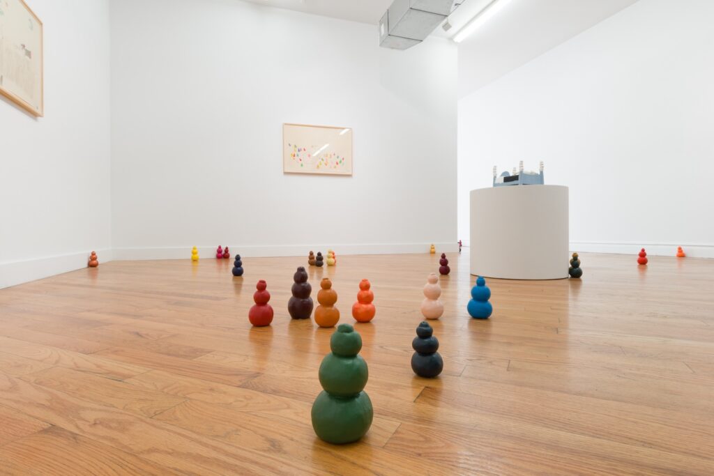 "Kong Play" is a sculptural installation consisting of 100 ceramic Kong sculptures that are installed throughout the gallery, mostly on the floor. A Kong is made up of three stacked spheres, with the largest on the bottom and the smallest on the top, they are reminiscent of a snowman. The bottom of each Kong is flat, enabling them to stand upright. Each Kong is painted with a different acrylic color. In total, there are 50 different colors, or 2 Kongs per color. In this installation shot, brightly colored Kongs are scattered across the floor. This photo was taken from a low vantage point and the single circular plinth in the mid ground towers above the Kongs. Two framed drawings hang on the wall.