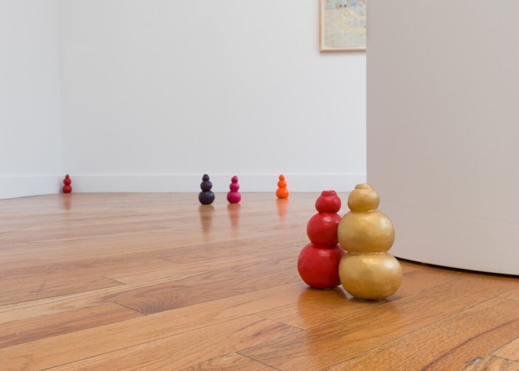 Installation view of "Kong Play", taken from floor level and focused close in on a pair of Kongs, one bright red, the other a shiny gold, that are nestled together. A few more colorful Kongs are scattered across the floor behind them and the bottom corner of a framed drawings peeks in from the upper righthand corner.