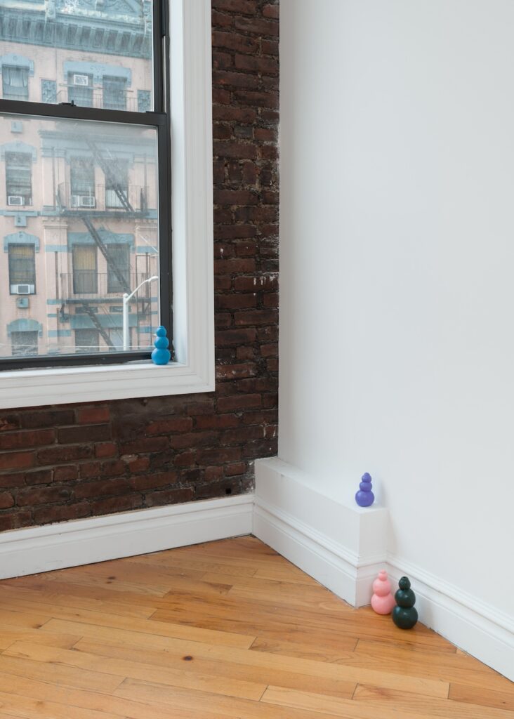 Vertical installation shot of "Kong Play". Four Kongs are installed in the front corner of the gallery, one on the windowsill, another on top of a bit of protruding moulding, while the last two are nestled together nearby on the floor. The wall on the left is made up of exposed brownish red bricks, the wall on the right is painted a bright white.