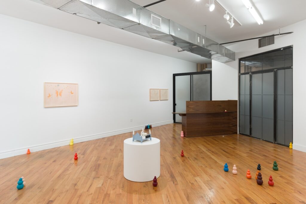 Installation view of "Kong Play", facing towards the closed front door of the gallery on the right and the darkly stained wooden desk on the left. In the center of the image, "Moon Bed" sits on a circular white plinth. Colorful Kongs cover the floor around it and three framed drawings hang on the wall behind, “Angel Dogs Flying to the Moon” by itself while the other two drawings are too far back to clearly make out.
