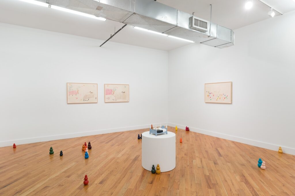 Installation shot of "Kong Play", centered on a white circular plinth with "Moon Bed" installed on top of it. Brightly colored Kongs are arranged across the floor around the plinth and there are three framed drawings hanging on the walls at the back, “Playing in Bed (with lobster)” and "Wild Girls" are paired together on one wall while "Pleasure Palace" is installed by itself on the other.