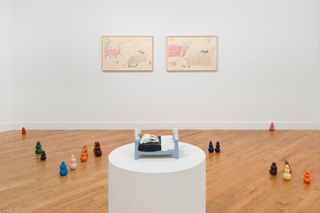 Installation shot of "Kong Play", in the foreground, "Moon Bed" sits on a white plinth, surround by a sea of Kongs. On the wall directly behind the plinth, two framed drawings are installed. On the left is “Playing in Bed (with lobster)” and on the right is "Wild Girls".