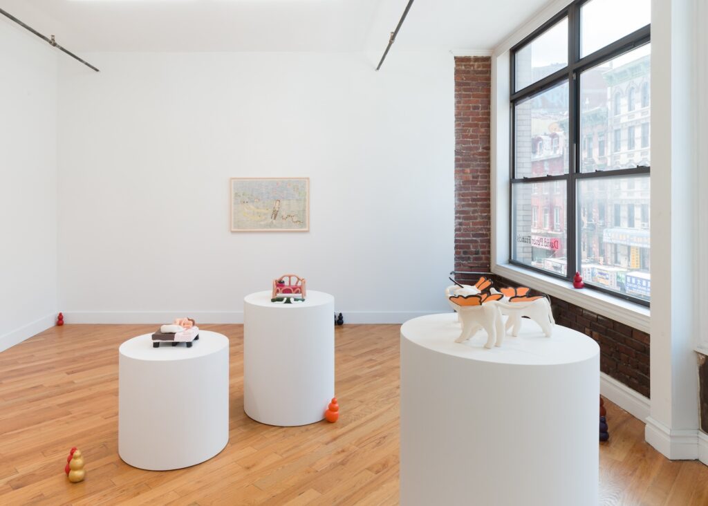 Installation shot of "Kong Play", taken in the back room of the gallery. The foreground is dominated by three circular white plinths of varying heights, the two on the left each have a ceramic bed sculpture on them while the plinth on the right has the three "Butterflydogs" on it. Kongs are scattered across the floor in pairs and alone. The drawing “In My Dreams, The Mississippi Carries Me” is hanging on the back wall.