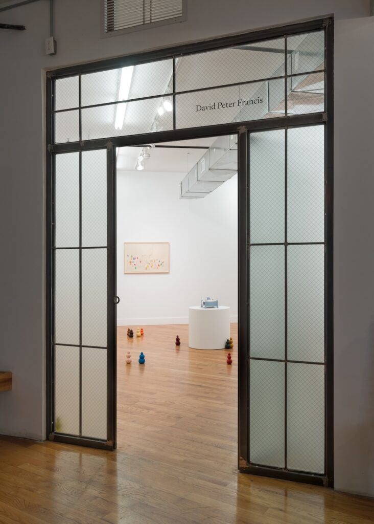 Vertical installation shot of "Kong Play" viewed from the lobby outside the gallery. The doorway is ringed by panes of frosted glass and the door is open. Beyond the door, Kongs are scattered across the floor, some in pairs, others alone. A single circular white plinth is topped by a blue bed sculpture. There is a single drawing hanging on the back wall.