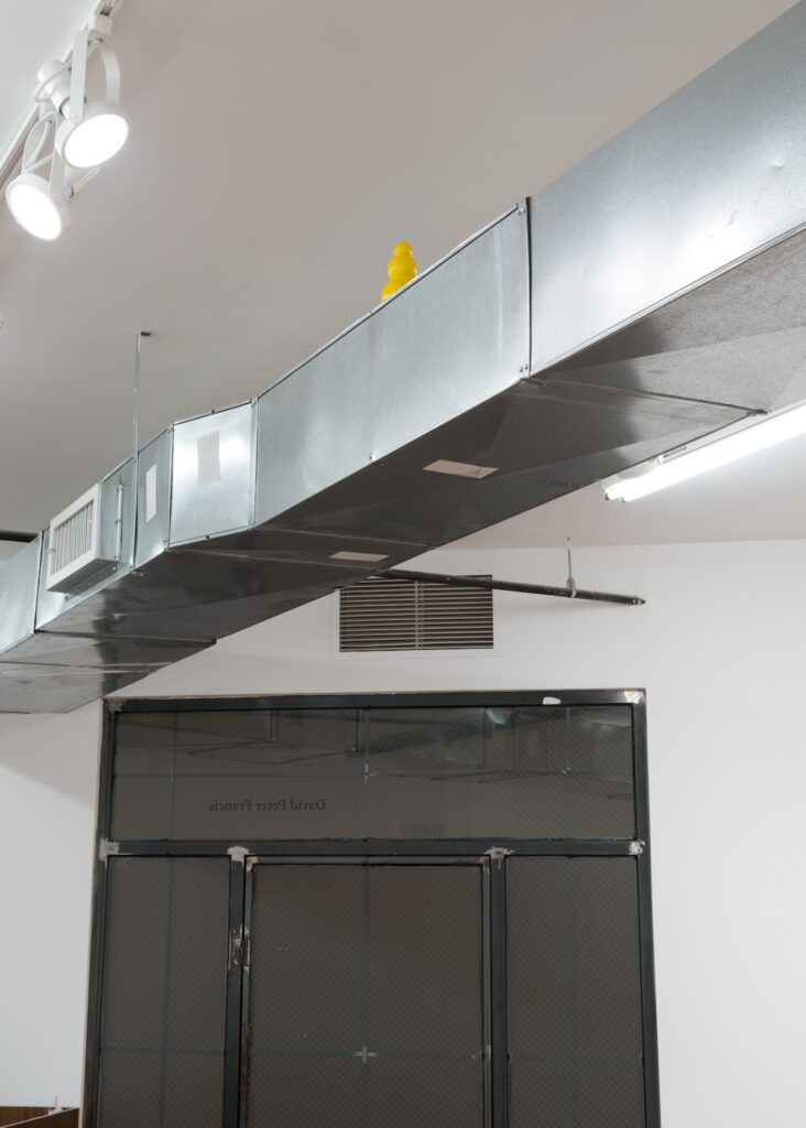 Vertical installation shot of "Kong Play" focused on a single, bright yellow Kong that has been placed, half-hidden, on top of the shiny silver ventilation duct. Only the top two spheres are fully visible.