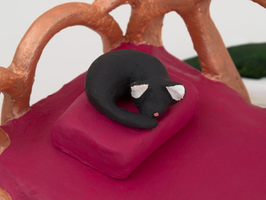 In this closeup of “Louisiana Day Bed”, the focus is on the black cat which is curled up asleep on the pillow. The white of its ears along with a tiny dot of pink on its nose are both highlighted.