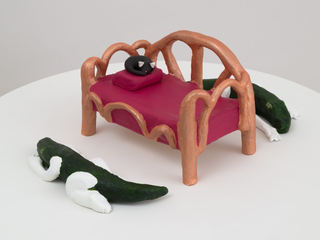 “Louisiana Day Bed” is a small, handmade ceramic sculpture featuring a day bed and two Alligatorgirls, that is alligators with human arms and legs. The bed has a rounded, arching headboard and footboard, both painted in a metallic copper color. The frame is chunky and uneven, giving it a playful, childlike appearance. The mattress is a solid reddish-pink color, with the same color extending down the sides, and the edges of the bed have wavy, scalloped detailing in copper. On top of the bed, near the headboard, is a small black cat curled up and asleep on a rectangular pink pillow. The cat has a pink dot for a nose and white ears. In front of the bed, on the white surface, lies an Alligatorgirl. The alligator’s body is dark green, with its arms and legs painted white. It is stretched out on its side, with its head resting on the ground and its legs extended, as if in a relaxed, sleeping pose. Another Alligatorgirl is partly visible behind the bed.
