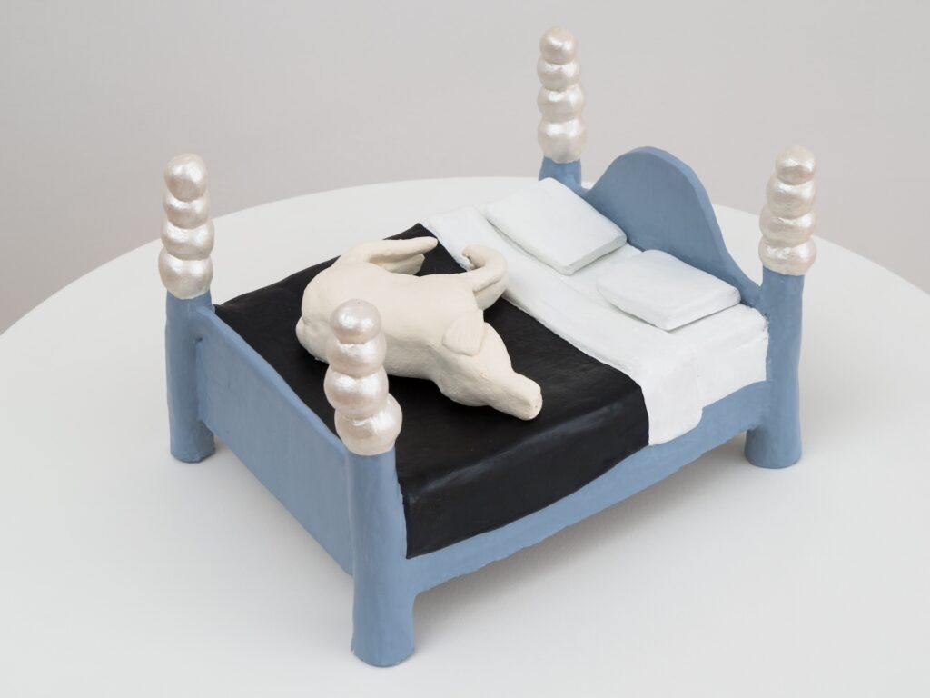 "Moon Bed" is a small, handmade ceramic sculpture of a bed with a dog resting on top of it. The bed is rectangular with four thick, rounded legs at each corner. Each leg extends upward into a tall post, and each post is topped with three large, shiny, pearl-like spheres stacked vertically. The posts and the bed frame are painted a soft, matte blue. The bed has a high, half moon shaped headboard, that is the same blue. The bedding consists of a dark, midnight blue blanket that covers most of the bed’s surface, with a white sheet draped over the top edge and hanging down the right side. Two white pillows are placed side by side at the head of the bed. On top of the blanket, is a white English Labrador Retriever lying on its side, curled up as if sleeping. Its legs are tucked in, and its tail curves around its body.