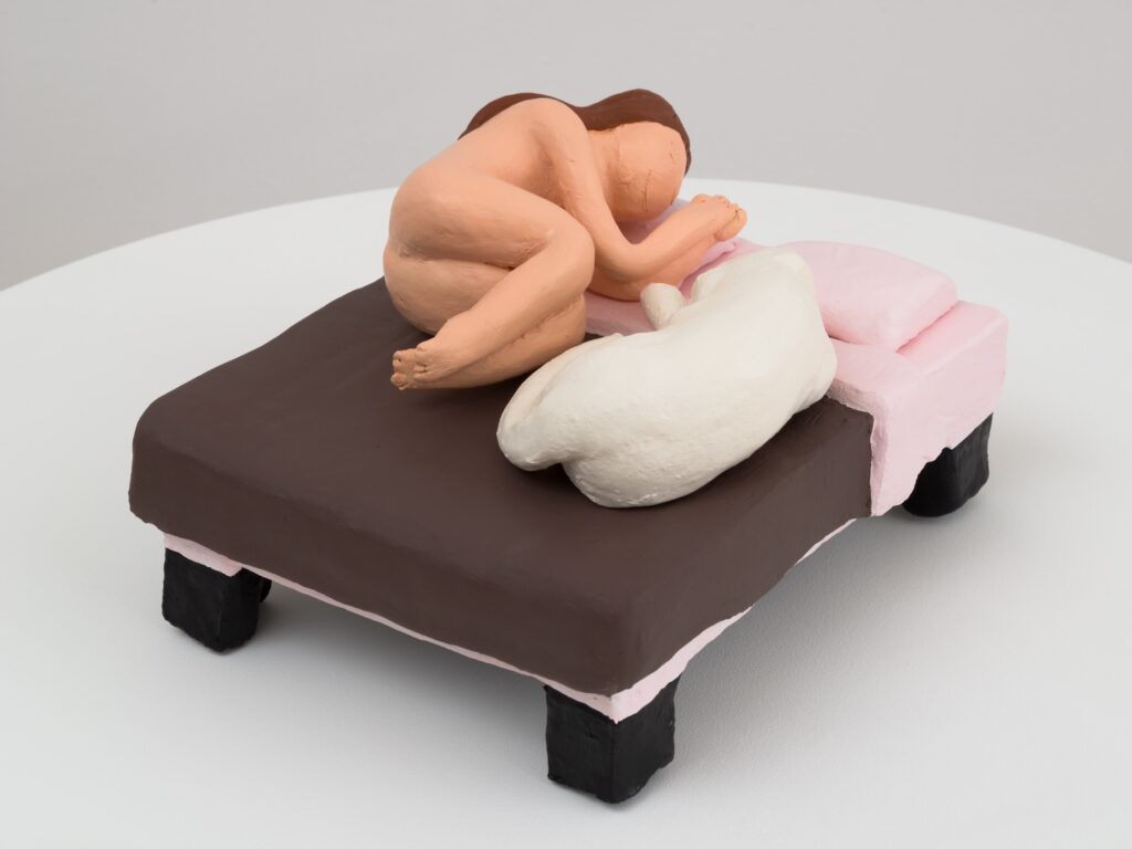 "Our Bed" is a small, hand built ceramic sculpture showing a cozy scene on a rectangular bed. The bed has a thick, chocolate-brown blanket covering the top, with a pink bed frame and two pink pillows at the head. The bed stands on four chunky, black legs. On the bed, a nude human figure with light peach skin and brown hair is curled up in a fetal position, lying on their left side. Their knees are tucked close to their chest, arms bent and hands together near their face, which is turned slightly downward. The figure’s facial features are minimal, with only the suggestion of a nose and closed eyes. Next to the person, a white dog is also curled up, mirroring the human’s position. The dog’s body forms a loose, circular shape, with its head tucked in and tail wrapped around. The sculpture has a handmade, almost cartoonish quality, emphasizing warmth, intimacy, and the bond between the person and the dog. The scene feels peaceful and safe, as if capturing a quiet moment of rest and companionship.