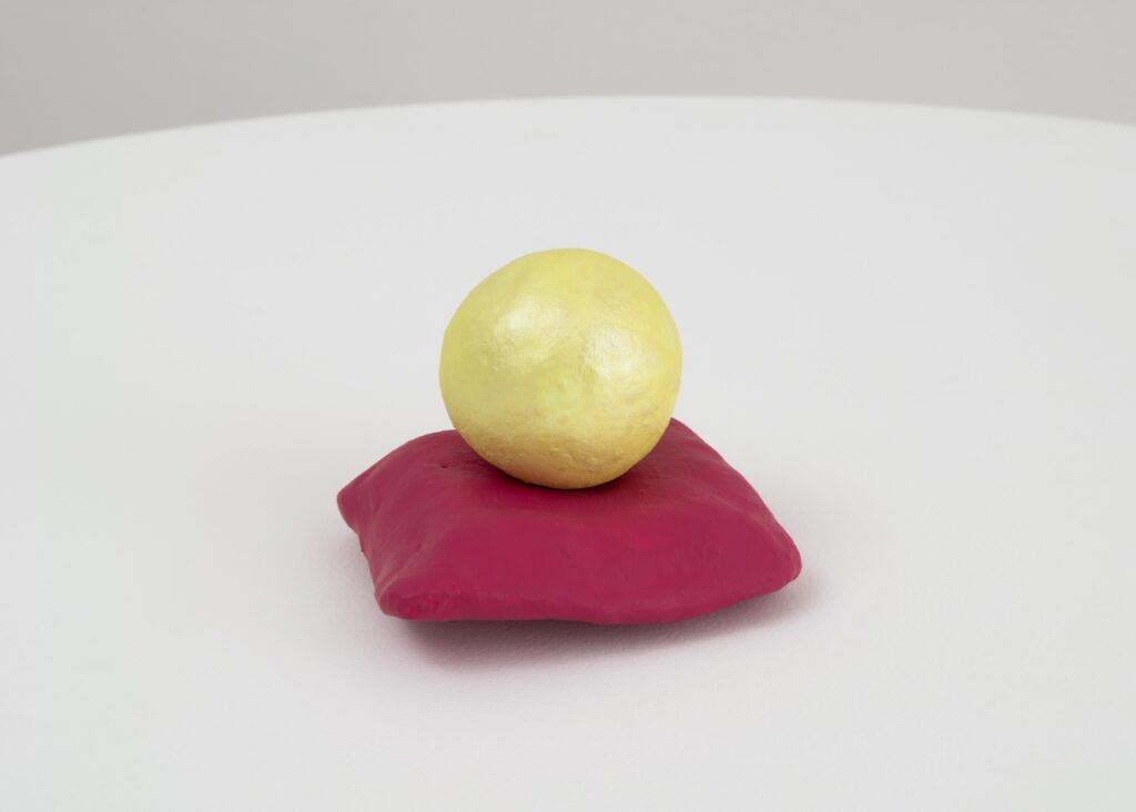“When I was a Ball of Light”, is a small abstract sculpture consisting of two main parts: a base and a sphere. The base resembles a soft, slightly squished magenta pillow, with rounded edges and a somewhat uneven surface. On top of the pillow, sits a single sphere, about the same width as the cushion. The sphere is a pale, buttery yellow with a subtle sheen, giving it a slightly reflective, almost glittery affect, as if it is glowing from within.