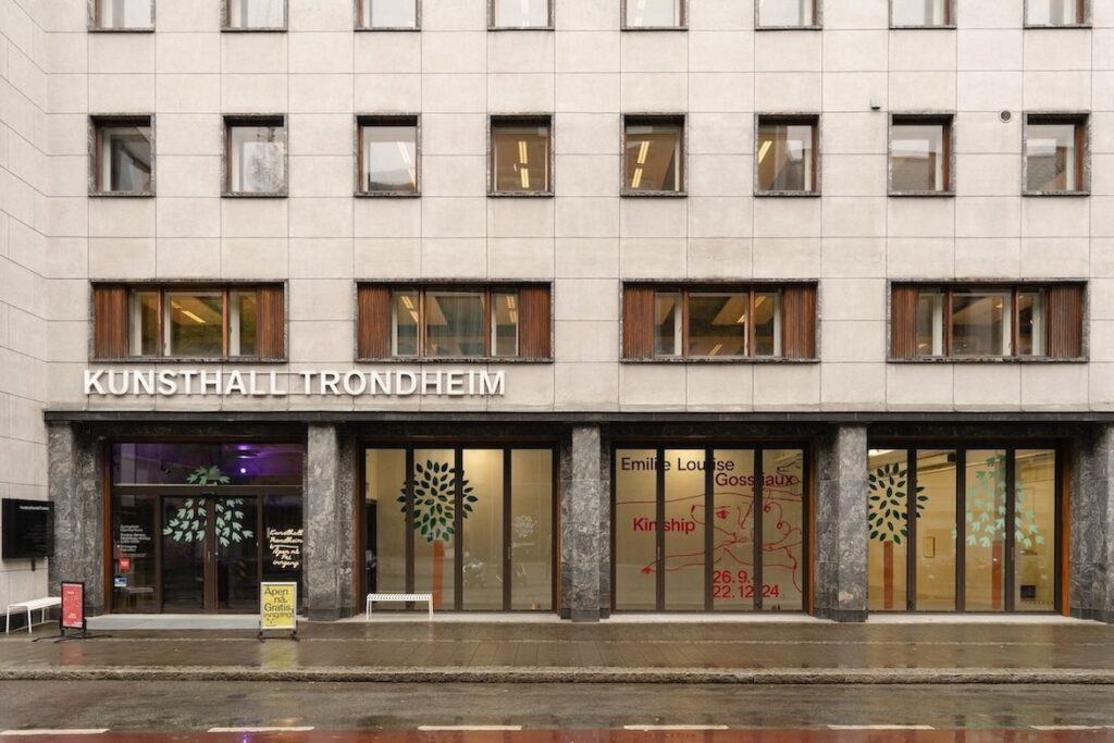 Exterior shot of Kunsthall Trondheim, which is a blocky grey building. The bottom floor is dominated by floor-to-ceiling windows which have colored pieces of vinyl attached to them that resemble the trees found in "White Cane Maypole Dance" at the Queens Museum. Through one window the words Emilie Louise Gossiaux and Kinship are visible, along with a red colored line drawing of a butterflydog.